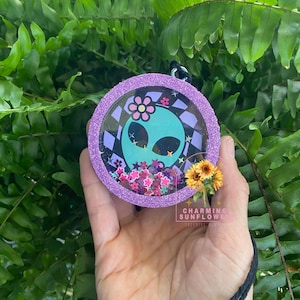 May include: A circular purple glitter ornament featuring a teal alien face graphic with a pink flower on its head. The center contains loose pink and purple flower-shaped sequins. A small sunflower charm is attached to the side of the frame.