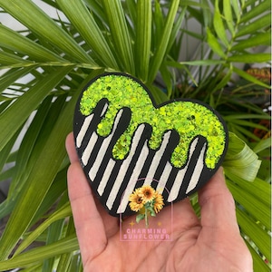 May include: A heart-shaped decoration with a black and white striped base, topped with dripping neon green glitter. The decoration has the text "Charming Sunflower" and a sunflower graphic.