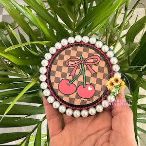 May include: Round coaster with a cherry design. The coaster features a brown and tan checkered background, a red glitter border, and a pearl bead edge. The cherry design has a pink bow and green stems. A small bouquet of sunflowers is visible.