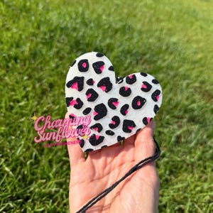 May include: A heart-shaped air freshener featuring a white base with a black and hot pink leopard print pattern. The surface has a textured, glittery appearance. A thin black string is attached to the top for hanging purposes.