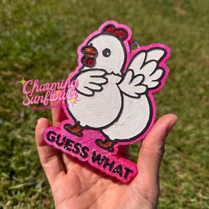 May include: A white chicken-shaped air freshener with a glittery pink border and black outline. The chicken has a red comb and orange feet. The text GUESS WHAT is printed in black glittery letters at the bottom of the pink base.