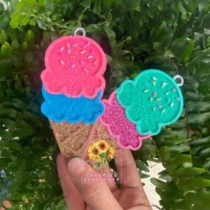 May include: Two glittery ice cream cone ornaments. One has pink and blue scoops with sprinkles, the other has pink and green scoops with sprinkles. The cones are brown. The text "CHARMING SUNFLOWER" is visible.