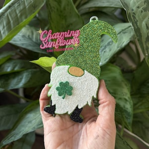 May include: A green glitter gnome car air freshener with a white beard, a shamrock, and black boots. The gnome has a tan nose and a green glitter hat. The text "Charming Sunflower CAR FRESHENERS" is visible in the upper left corner.