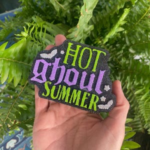 May include: A black glittery cutout sign featuring the text "HOT ghoul SUMMER" in lime green and purple glitter lettering. The design includes small silver glitter bat accents positioned around the text on a dark, textured, sparkling background.