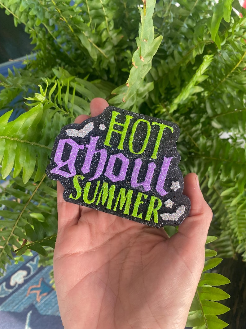 May include: A black glittery cutout sign featuring the text "HOT ghoul SUMMER" in lime green and purple glitter lettering. The design includes small silver glitter bat accents positioned around the text on a dark, textured, sparkling background.