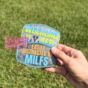 May include: A two-part glittery car air freshener with a gray textured surface. The top section features the text WEEKEND HIKER in lime green and blue. The bottom section features the text EASILY DISTRACTED BY MILFS in orange and light blue.