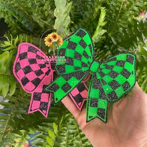 May include: A pair of glittery hair bows featuring a checkered pattern. One bow is bright neon pink and black, while the other is vibrant neon green and black. Both bows have a textured, sparkling finish and are shaped with multiple loops.