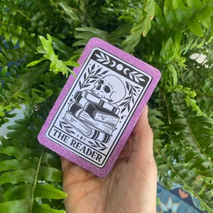 May include: A rectangular card with a glittery purple border featuring a black and white illustration of a human skull resting on a stack of three books. The text THE READER is printed in a white rectangular box at the bottom of the card design.