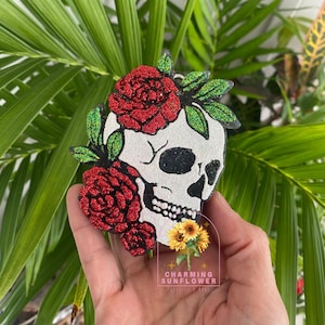 May include: A white skull-shaped ornament with red glitter roses and green glitter leaves. The skull has black eye sockets and a black nose. The ornament has the text "Charming Sunflower" and a sunflower graphic.