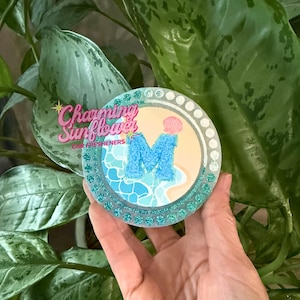 May include: A round car air freshener with a beach-themed design. The center features a light blue letter "M" on a light blue and white background, with a pink seashell. The border is teal glitter, and the text "Charming Sunflower CAR FRESHENERS" is visible.