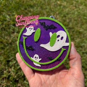 May include: A circular purple glitter car freshener featuring a green smiley face outline, two green oval eyes, and three white ghost figures. The design includes small black glitter bats scattered across the surface. The text Charming Sunflower Car Fresheners is printed in pink and white in the upper left corner.