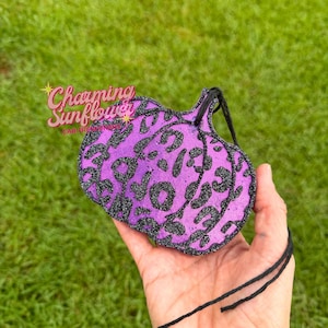 May include: A pumpkin-shaped air freshener featuring a vibrant purple base color with a black glitter leopard print pattern. The object includes a black string attached to the top stem for hanging purposes and displays a logo in the upper left.
