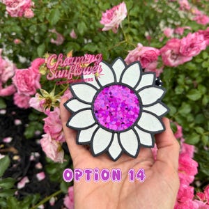 May include: A flower-shaped car air freshener featuring white petals with black outlines and a center filled with bright pink glitter. The text "Charming Sunflower" is printed in pink above the object, and "OPTION 14" is printed in pink below.