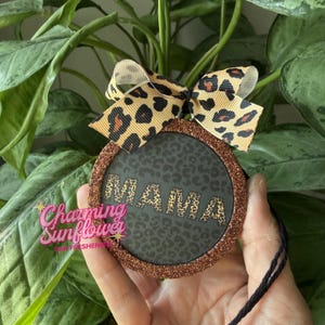 May include: Round car air freshener with a leopard print design and the word "MAMA". The air freshener has a glittery bronze border and a leopard print bow. The text "Charming Sunflower" is also visible.