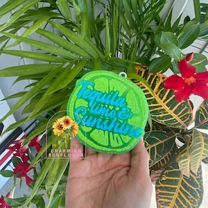 May include: A circular lime-shaped ornament with a textured green glitter finish. The text "Tequila lime & Sunshine" is embossed in bright blue glitter across the center. A small metal loop is attached to the top for hanging purposes.