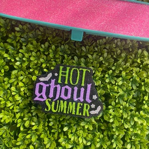 May include: A rectangular rearview mirror cover with a bright pink glitter finish and a teal frame. Below it is a black, glittery, die-cut sign featuring the text "HOT ghoul SUMMER" in lime green and purple, accented with small silver glitter bats.