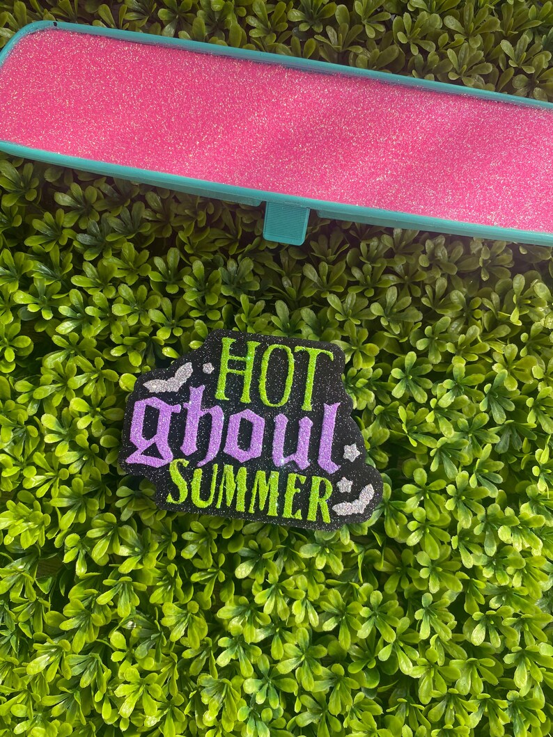 May include: A rectangular rearview mirror cover with a bright pink glitter finish and a teal frame. Below it is a black, glittery, die-cut sign featuring the text "HOT ghoul SUMMER" in lime green and purple, accented with small silver glitter bats.