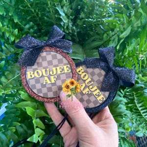 May include: Two round ornaments with the text "BOUJEE AF" in yellow, set against a brown and tan checkered background. Each ornament has a glittery brown border and a black glitter bow. A small bouquet of sunflowers is held in front of the ornaments.