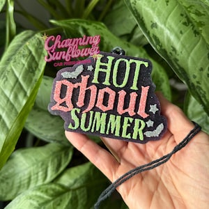 May include: A black car air freshener with glittery text that reads HOT GHOUL SUMMER. The word HOT and SUMMER are in lime green, while the word ghoul is in pink. The design features small silver glittery bats and stars on a black background.