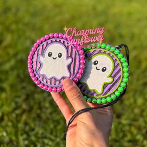 May include: Two circular hanging ornaments featuring a white ghost graphic on a striped background. One ornament has a pink beaded border, and the other has a green beaded border. The text Charming Sunflower is visible above the ornaments.