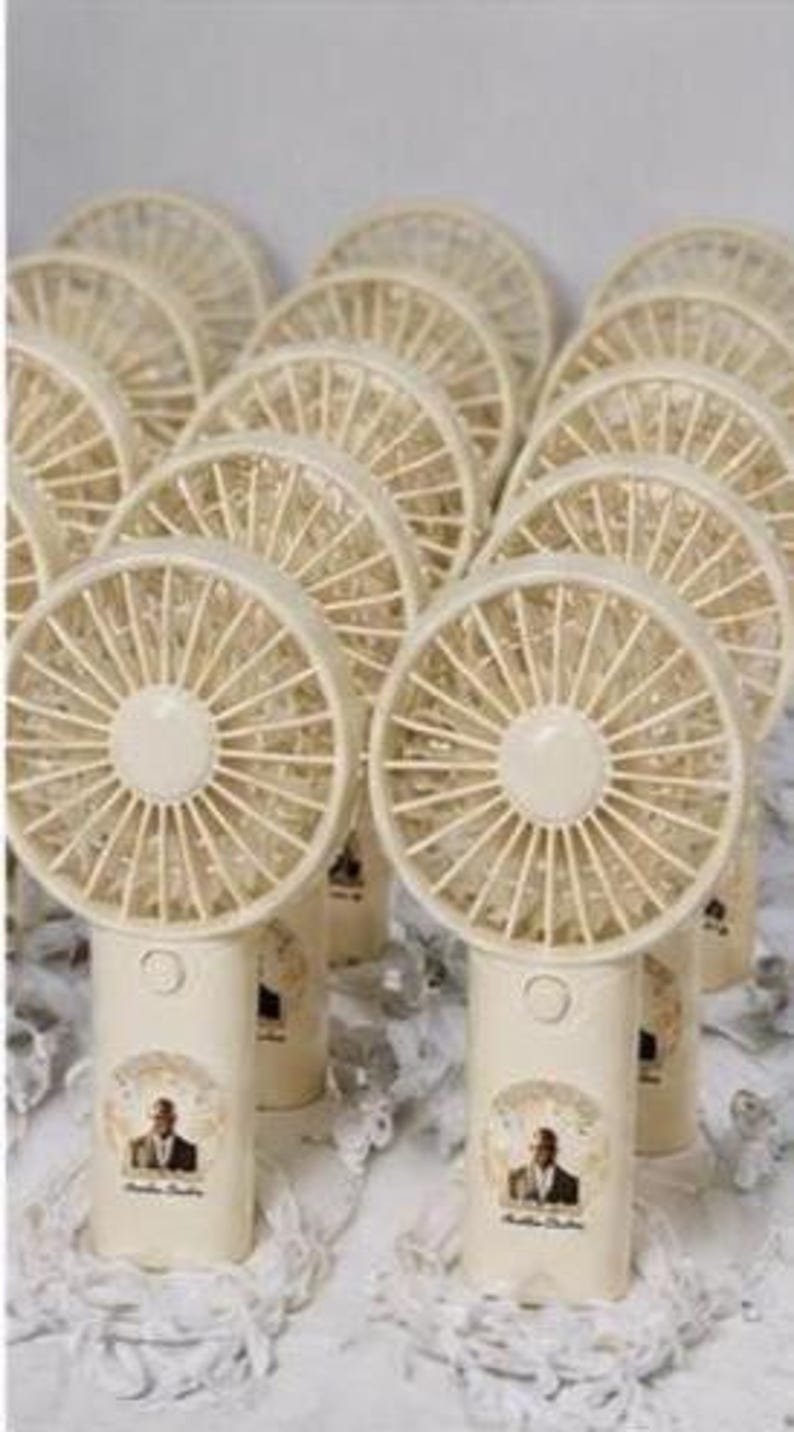 May include: A collection of cream-colored portable fans with a circular fan blade design. Each fan has a cylindrical base with a small image of a person. The fans are arranged on a white surface.