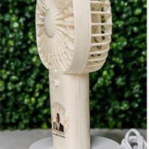 May include: A cream-colored portable fan with a round head and a handle, resting on a white circular base. The fan has a protective cage and a small sticker with a person's image. A white USB cable and plug are visible.
