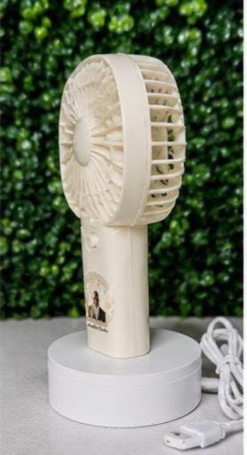 May include: A cream-colored portable fan with a round head and a handle, resting on a white circular base. The fan has a protective cage and a small sticker with a person's image. A white USB cable and plug are visible.
