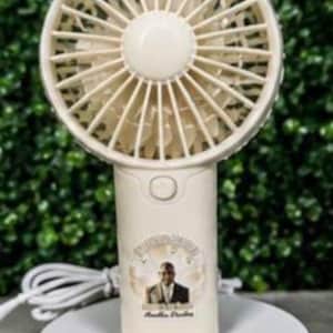 May include: A cream-colored handheld fan with a round fan head and a cylindrical handle. The fan is on a white rotating base. The handle has a button and a circular sticker with a person's image and text.