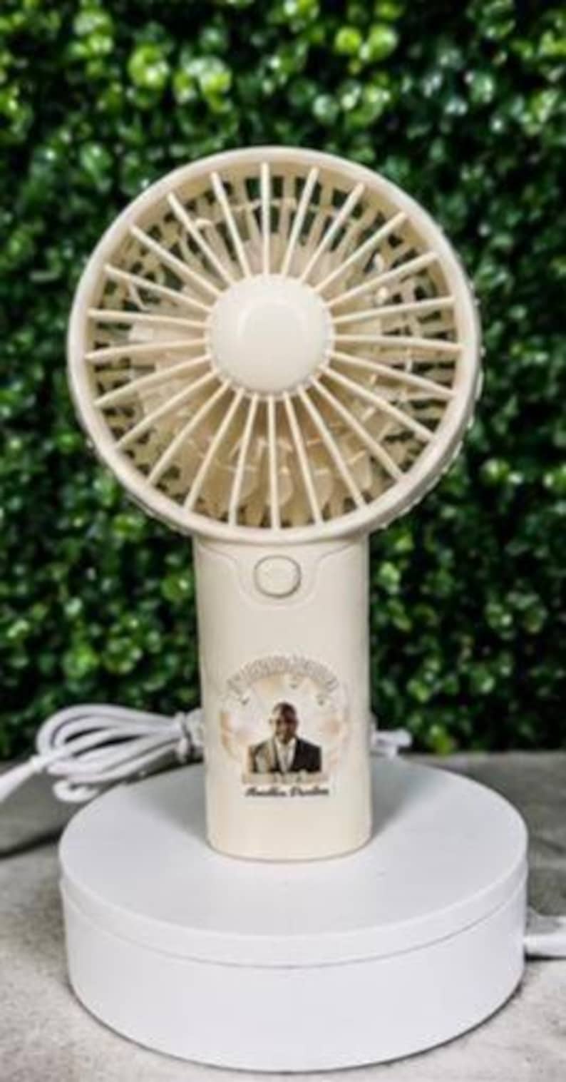 May include: A cream-colored handheld fan with a round fan head and a cylindrical handle. The fan is on a white rotating base. The handle has a button and a circular sticker with a person's image and text.