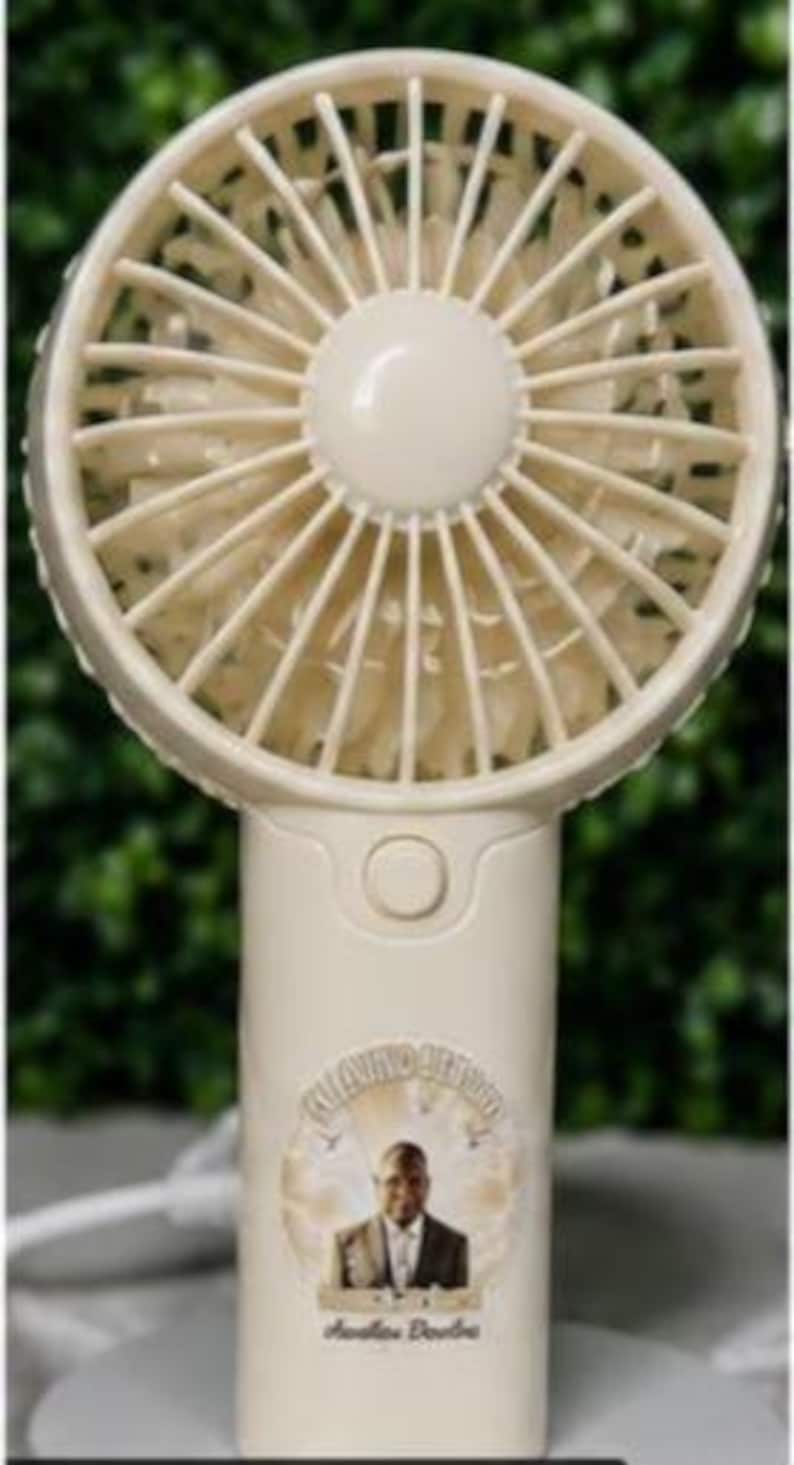 May include: A cream-colored handheld fan with a circular fan head and a cylindrical handle. The fan head has multiple blades. The handle features a button and a circular image of a person with text. The fan is on a white surface.