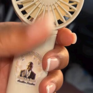 May include: A white and beige portable fan is held in a hand. The fan has a sticker with a photo and text. The fan is battery-operated and designed for personal cooling.