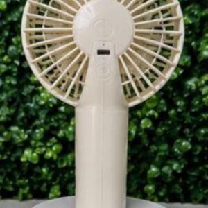 May include: A cream-colored portable fan with a round head and a cylindrical handle. The fan is mounted on a white, circular base. The fan's blades are visible, and there is a small digital display on the handle.