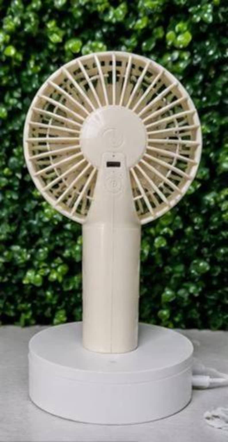 May include: A cream-colored portable fan with a round head and a cylindrical handle. The fan is mounted on a white, circular base. The fan's blades are visible, and there is a small digital display on the handle.