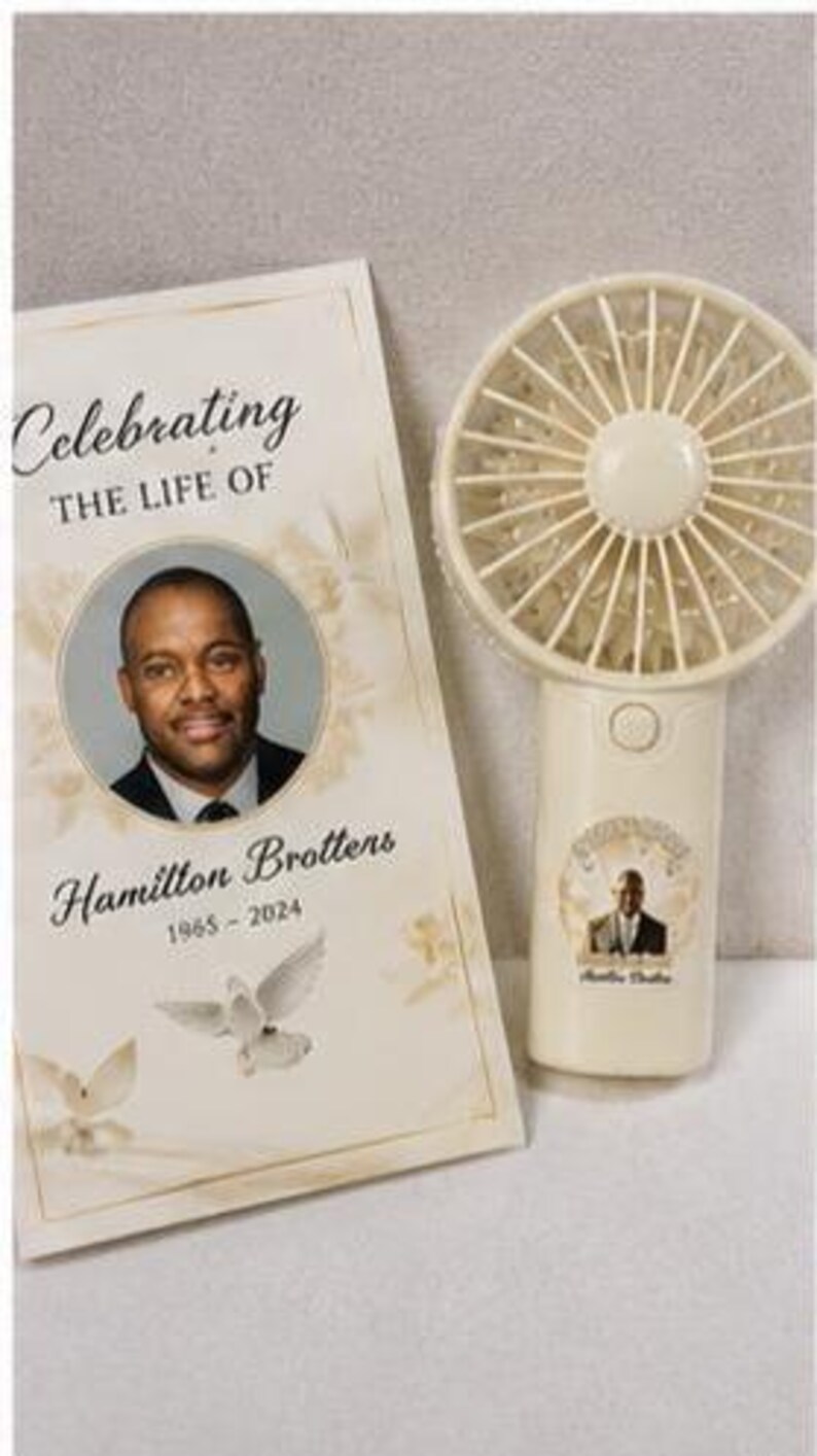 May include: A cream-colored handheld fan and a memorial card. The card reads "Celebrating The Life Of Hamilton Brottens 1965-2024" with a photo and dove graphic. The fan has a matching photo and is off-white.