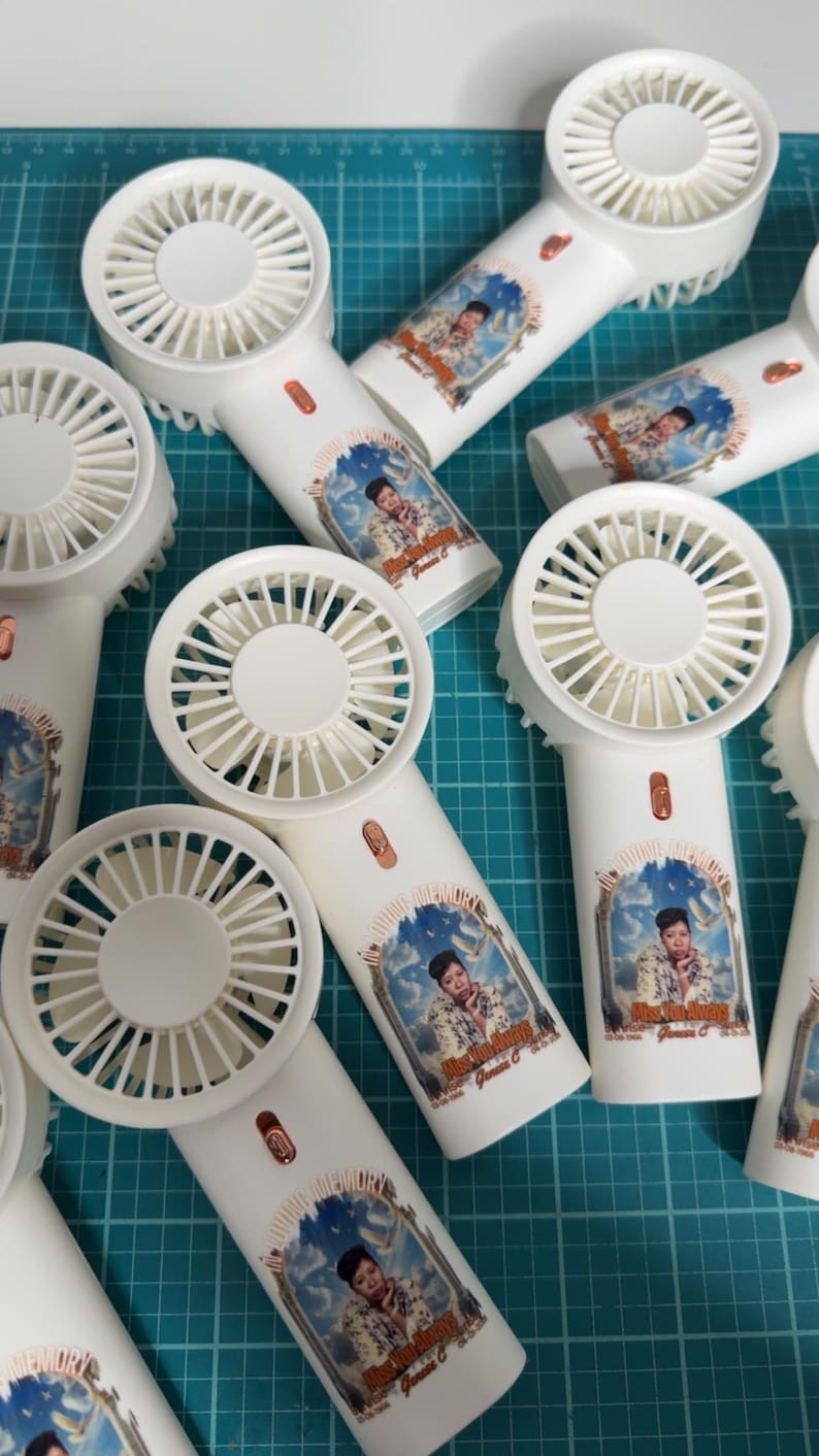 May include: A collection of white portable fans with a circular fan head and a handle. Each fan has a printed image of a person with text. The fans are arranged on a blue grid surface.