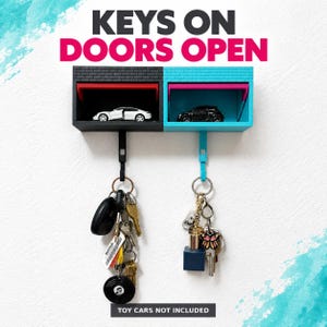 May include: A wall-mounted key holder featuring two garage-style compartments, one black and one turquoise, with brick-textured facades. Each garage has a hook underneath holding a set of keys. The text KEYS ON DOORS OPEN is displayed above.