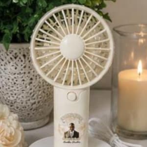 May include: A cream-colored, handheld electric fan with a round head and a handle. The handle has a small image of a man in a suit and the words "Amelia Brothers". The fan is on a white circular stand. A white cord extends from the base.