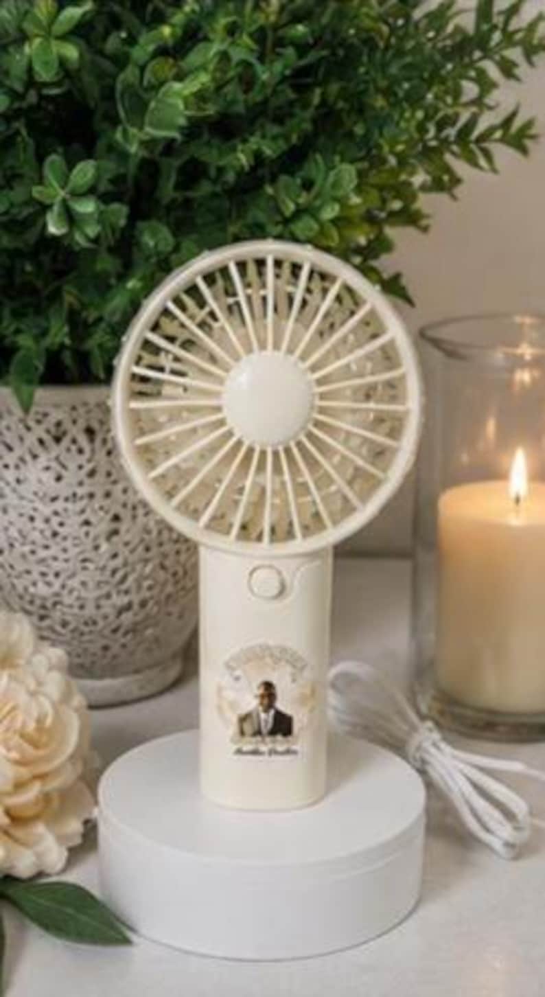 May include: A cream-colored, handheld electric fan with a round head and a handle. The handle has a small image of a man in a suit and the words "Amelia Brothers". The fan is on a white circular stand. A white cord extends from the base.