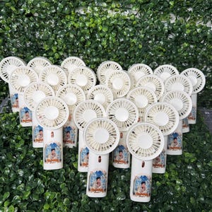 May include: A collection of white handheld fans with a portrait on the handle. The fans have a circular fan blade design and are arranged in a group against a backdrop of green foliage.