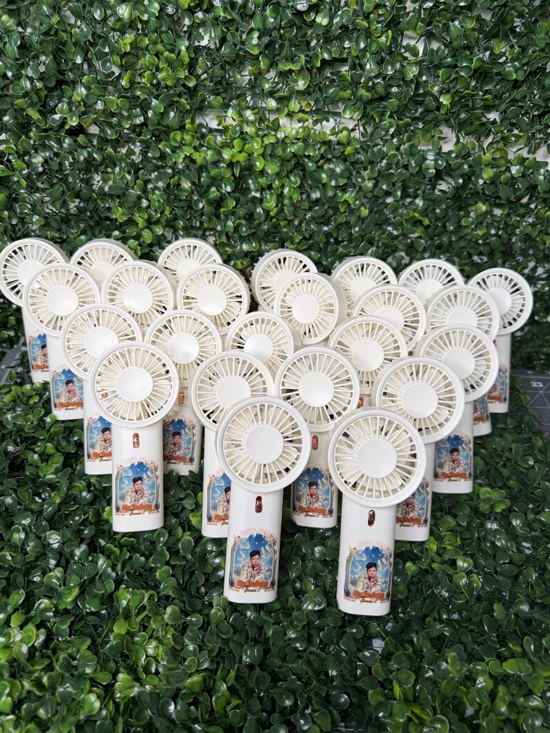 May include: A collection of white handheld fans with a portrait on the handle. The fans have a circular fan blade design and are arranged in a group against a backdrop of green foliage.
