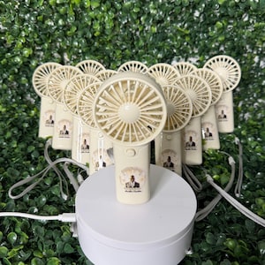 May include: A collection of beige handheld fans with a circular fan blade design. Each fan has a small image on the handle and a gray lanyard. The fans are arranged on a white platform against a backdrop of green foliage.