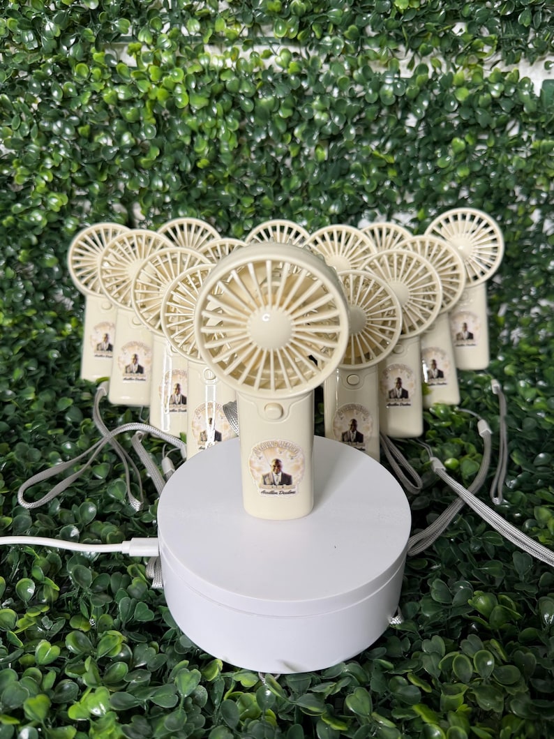 May include: A collection of beige handheld fans with a circular fan blade design. Each fan has a small image on the handle and a gray lanyard. The fans are arranged on a white platform against a backdrop of green foliage.