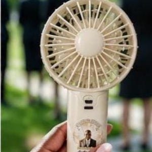 May include: A cream-colored, handheld personal fan with a circular fan head and a handle. The handle features a small portrait and text. The fan is held in front of a blurred background of people.
