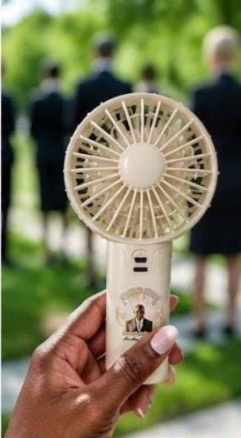 May include: A cream-colored, handheld personal fan with a circular fan head and a handle. The handle features a small portrait and text. The fan is held in front of a blurred background of people.