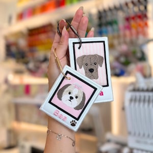 May include: Two rectangular embroidered keychains featuring dog portraits. One keychain displays a brown dog on a pink striped background with a black border. The other shows a white and brown dog with the name Bea and a paw print embroidered in pink.