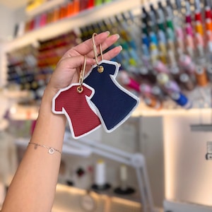 May include: Two fabric keychains shaped like t-shirts, one in deep red and one in navy blue, each featuring a white stitched border and a gold-toned metal carabiner clip attached through a reinforced eyelet at the neckline of the garment shape.