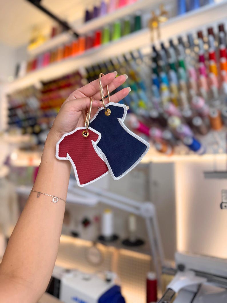 May include: Two fabric keychains shaped like t-shirts, one in deep red and one in navy blue, each featuring a white stitched border and a gold-toned metal carabiner clip attached through a reinforced eyelet at the neckline of the garment shape.