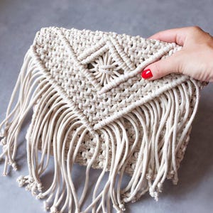 Puede incluir: Un bolso de mano tipo sobre en macramé color blanco roto con una solapa triangular y un patrón central de nudos en forma de diamante. El cuerpo del bolso está hecho de cordón grueso anudado y tiene largos flecos colgantes en el borde.