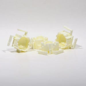 Puede incluir: Un conjunto de tres componentes de plástico color blanco hueso que presentan un centro cilíndrico hueco de múltiples facetas con brazos rectangulares salientes y soportes estructurales. Estas piezas pequeñas y rígidas tienen un acabado mate.