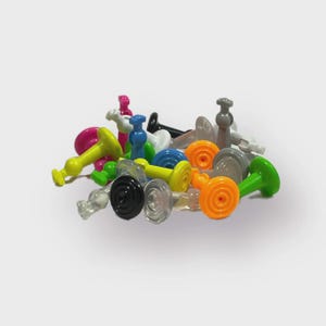 Puede incluir: Una colección de pequeñas chinchetas o piezas de juego de plástico multicolor. El surtido incluye tonos vibrantes de amarillo, naranja, verde, azul, rosa, negro, blanco, gris y piezas transparentes, cada una con una base circular plana.